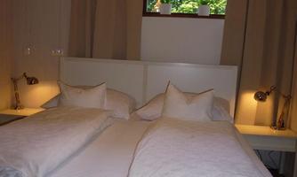 A cozy bedroom with a double bed and light sheets. The window shows green plants in the background.