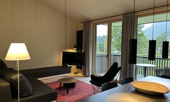A modern living room with a cozy couch and a dining area. Large windows offer a view of nature.