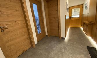 A bright hallway with a wooden door. On the left and right are several doors, and a window lets in daylight.