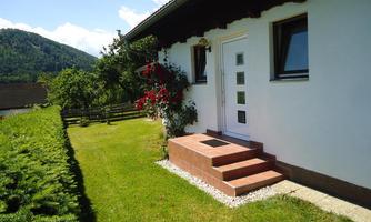 A beautiful house with an entrance staircase and a blooming garden. In the background, gentle hills and a clear sky can be seen.