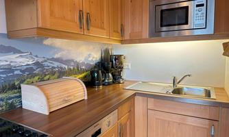 A modern kitchen with wooden furniture and a large window. In the background, a mountain motif can be seen, and there is a coffee machine as well as a microwave.