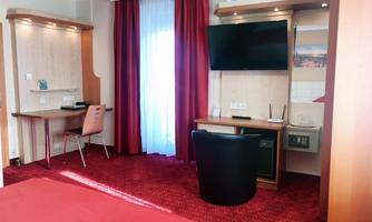 A modern hotel room with a red carpet and a large window. There is a desk, a television, and comfortable furniture.