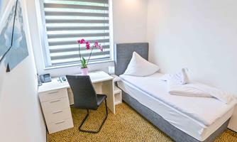 A simple room with a single bed, a desk, and a plant on the table. The walls are light and the window has blinds.