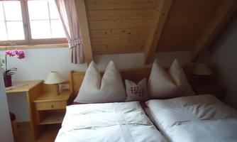 A cozy bedroom with two single beds and heart-shaped pillows. There is a window with curtains and a houseplant on the nightstand.