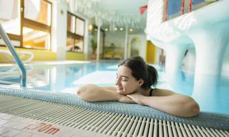 A woman relaxes at the edge of a swimming pool. In the background, brightly lit windows and colorful slides can be seen.