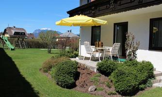 A sunny garden with a table and chairs under a yellow sun umbrella. In the background, a slide and the mountains are visible.