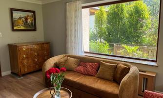 A cozy living room with a brown sofa and a coffee table. Through the large window, you can see a green garden with many plants.