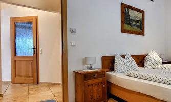 A cozy bedroom with a wooden bed and pillows. In the background, an open door is visible, showing an additional room.