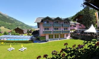 A colorful hotel with a large garden and a pool in the mountains. In the background, you can see green hills and a clear sky.