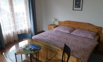 A cozy bedroom with a large bed and a dining table. Large windows provide plenty of daylight and an inviting atmosphere.