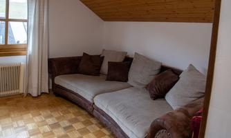 A cozy living room with a large brown sofa and many cushions. The room has wooden flooring and a window that lets in plenty of natural light.