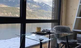 A modern workplace with a glass table and comfortable chairs overlooks a picturesque lake and the surrounding mountains. The large window front lets in plenty of daylight.