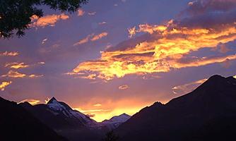 A beautiful sunset over the mountains, with glowing orange and purple clouds. The snow-capped peaks appear in the background.