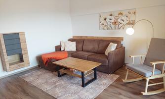 A cozy living room with a brown sofa, a wooden table, and an armchair. On the wall hangs a piece of art with floral motifs.