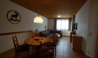 A cozy living area with a dining table and chairs. The room has large windows and a wooden ceiling.