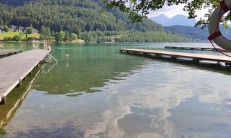 A quiet lake surrounded by mountains and trees. In the foreground, wooden walkways leading into the water are visible.