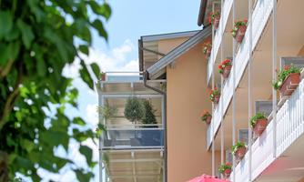 A modern building with balconies and flower boxes. In the foreground, there is a red sun umbrella under a tree.