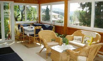 A cozy winter garden with rattan furniture and a table. Large windows provide a view of the green surroundings.