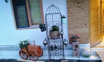 A decorative entrance with a rustic wooden cart and a metal plant column. Flowers in various pots give the area a cheerful atmosphere.