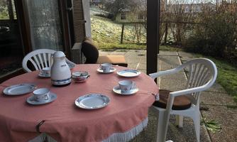 A set dining table on a terrace with dishes and a coffee pot. In the background, a green meadow and a clear sky are visible.