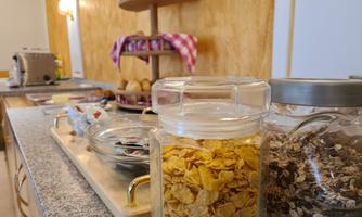 A breakfast table with various cereals in jars. In the background, rolls and a toaster can be seen.
