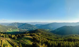 A breathtaking panoramic view of green hills and forests under a clear blue sky. The sun shines over the landscape, creating a peaceful atmosphere.