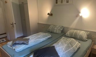 A cozy bedroom with two single beds and soft color tones. The walls are plain, and there is a closet nook as well as decorative wall lamps.