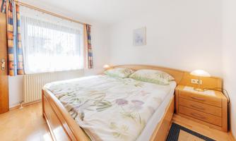 A bright bedroom with a large double bed and floral bedding. There is a dresser and curtains that let light in.