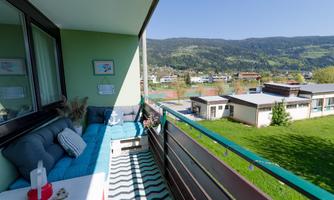 A cozy balcony with a seating area and a view of green hills and modern buildings. The sun is shining and the sky is clear.