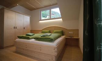 A bright bedroom with a large bed and two green pillows. There is a wooden wardrobe in the room and you can see windows overlooking greenery.
