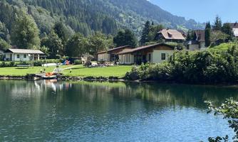 A peaceful landscape with a clear lake and beautiful green meadows. In the background, mountains and some cozy houses can be seen.