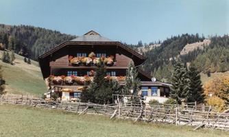 A beautiful wooden house with colorful flowers on the balcony. It stands in a green meadow, surrounded by mountains and trees.