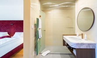 A modern bathroom with a glass shower and a large round mirror. In the background, you can see a stylish bedroom with a red bed.