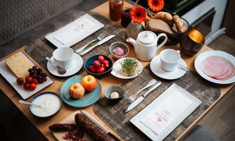 A beautifully set table with various breakfast options. There is cheese, cold cuts, fruit, yogurt, and rolls, as well as tea and jam.