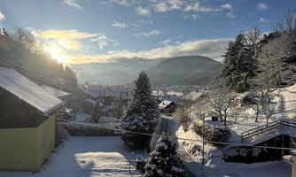 A winter landscape with snow-covered roofs and trees. The sun rises behind the mountain and illuminates the scene.