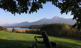 A person is riding a bicycle on a meadow with a view of mountains and a lake. The sky is clear and the landscape is green and peaceful.