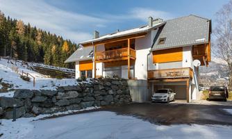 A modern house in winter with snow on the ground. The surroundings are surrounded by fir trees and the sky is clear.
