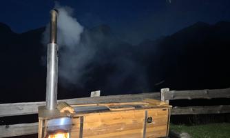 A wood-fired bath barrel stands outdoors, surrounded by mountains in the darkness. Smoke rises from the chimney.