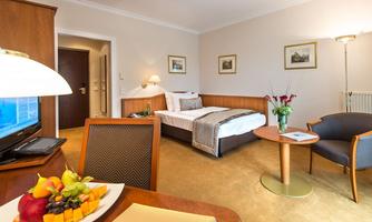A cozy hotel room with a double bed, a seating area, and a small table. On the table is a fruit bowl with fresh fruits.