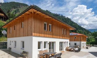 Two modern wooden houses in a picturesque mountain landscape. The sky is clear and the surroundings are green and inviting.