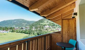 A balcony with a wooden railing and a table as well as a chair. In the background, green hills and a picturesque landscape can be seen.