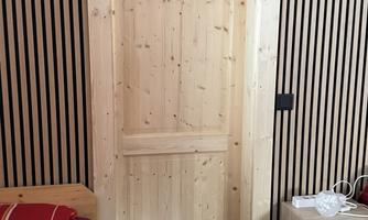 A bright wooden door in a modernly designed room. The wall is decorated with wooden slats, and there is a bed as well as a small nightstand.