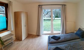 A simple, bright bedroom with two beds and a wardrobe. Large windows offer a view of the garden.