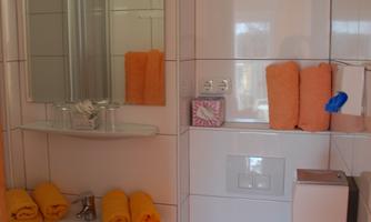 A modern bathroom with white tiles. Towels in orange and yellow hang decoratively, and a toilet is visible.