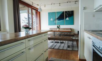 A modern kitchen with light wood and elegant cabinets. The living area is open, featuring pictures on the wall and a cozy dining table.