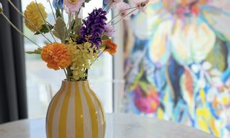 A colorful bouquet in a striped vase is on a table. In the background, a colorful floral painting can be seen.