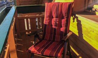 A comfortable chair with a red-striped cover is on a balcony. The sun is shining and casting shadows on the wooden floor.