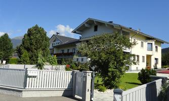A cozy residential house surrounded by trees and a well-maintained garden. The sun is shining in a clear blue sky.