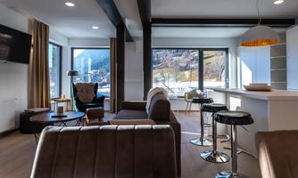 A modern living room with large windows and a view of the mountains. The decor is elegant and cozy with a sofa, chairs, and a dining area.