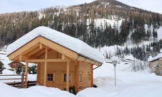 A wooden house with a snow-covered roof stands in a snowy landscape. Forested hills are visible in the background.